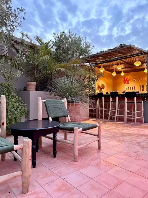 Terrasse sur le toit du Riad 191 Marrakech avec carrelage rose, table noire, bar et lanternes dorées sous ciel bleu