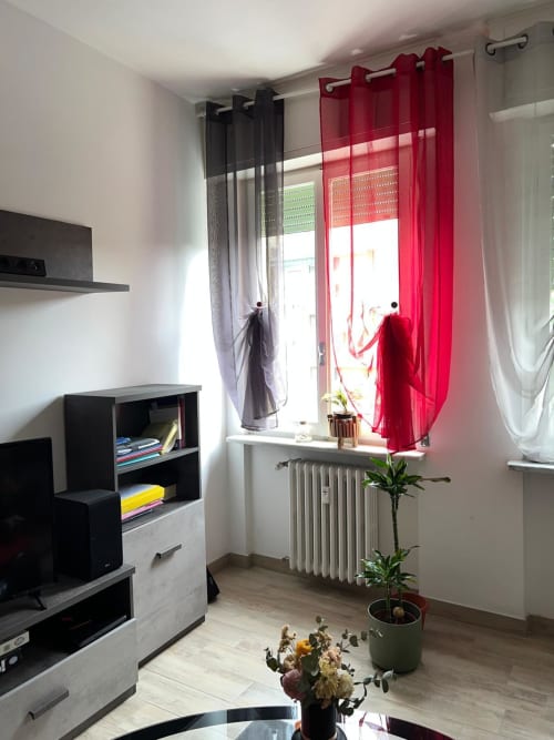 Luminoso soggiorno dell'appartamento MiSaNi Orchidea a Novara con tende rosse, scaffali, termosifone e piante in vaso