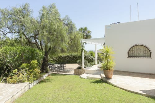 RB Algarve villa garden with manicured lawn, mature trees, modern white building, patio seating, and pergola in Boliqueime,
