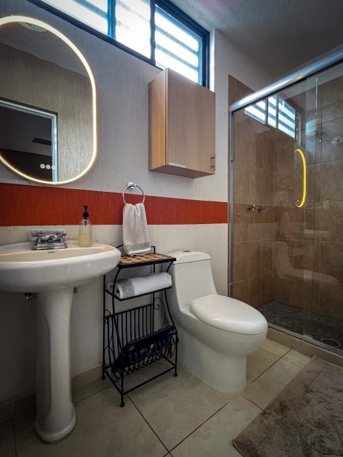 Baño moderno en apartamento Living UP Santiago de Querétaro con pared de acento rojo, espejo iluminado redondo, lavamanos pedestal, inodoro,