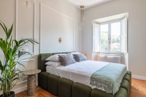 Camera da letto moderna in appartamento romano con letto rivestito in verde, biancheria bianca, pianta in vaso, pavimenti in legno e vista sulla città