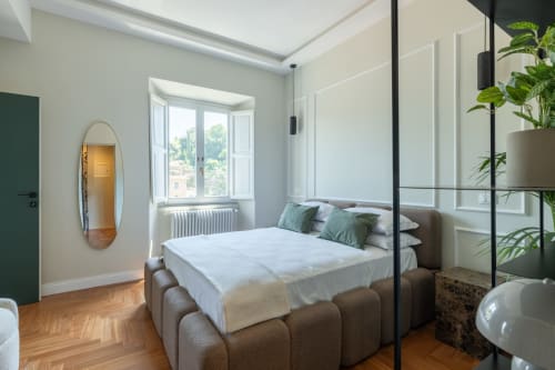Camera da letto moderna in appartamento romano con letto imbottito grigio, biancheria bianca, piante in vaso, specchio ovale, grandi finestre
