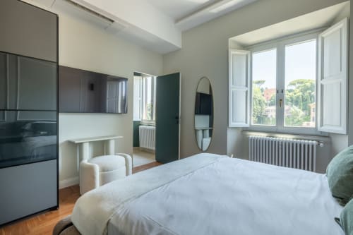 Camera da letto moderna in appartamento romano con letto bianco, accenti verdi, pavimento in legno, ampi affacci sulla città