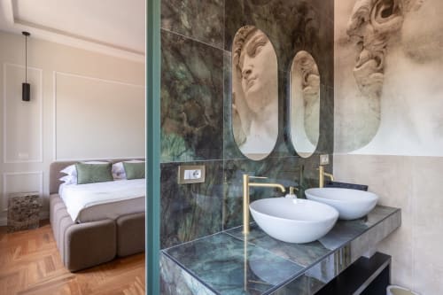Bagno moderno in appartamento romano con due lavabi bianchi, pareti in marmo verde, specchi ornamentali e vista sulla camera da letto