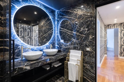 Lussuoso bagno di appartamento romano con pareti in marmo nero, specchi circolari illuminati in blu neon, lavabi bianchi e asciugamani dorati