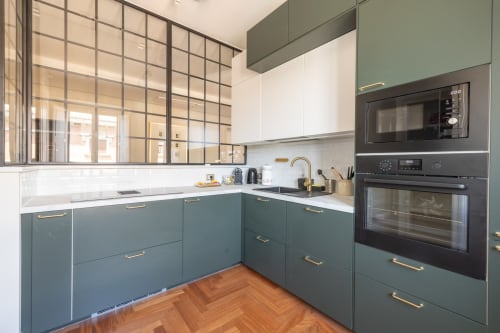 Cucina moderna in appartamento romano con mobili teal, top bianchi, maniglie dorate, finestra a quadretti e elettrodomestici neri.