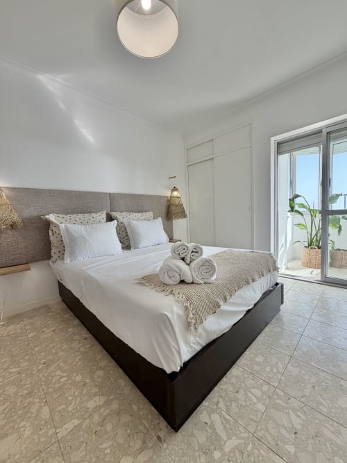Quarto moderno com vista para o mar no apartamento de Aroeira. Cama queen com cabeceira cinza, roupas de cama brancas, toalhas enroladas e decoração costeira