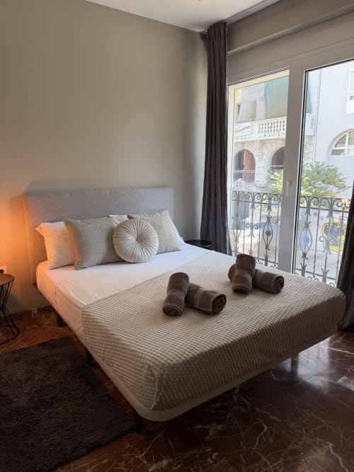 Modern bedroom in The Ruzafa Market Apartment, Valencia, with gray upholstered bed, city view balcony, and rolled towels