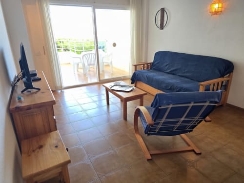 Salon lumineux avec canapé-lit bleu et fauteuil, bureau en bois, portes-fenêtres vue mer, sol carrelé à Rosas en bord de plage