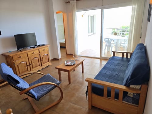 Lumineux séjour de l'appartement en bord de mer à Roses avec mobilier en bois, sièges bleus, TV et portes-fenêtres donnant sur la mer