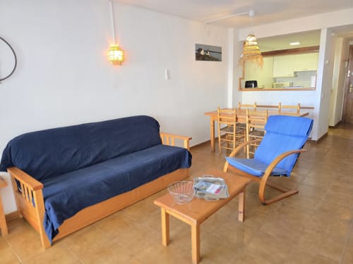 Spacieux salon dans un appartement en bord de mer à Roses avec mobilier en bois, sièges bleus et cuisine ouverte avec vue mer