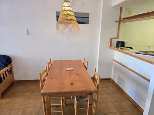 Coin repas dans appartement en bord de mer à Roses avec table en bois, chaises, lampe suspendue en tissage et kitchenette