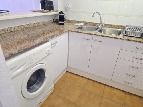 Cuisine moderne avec plan de travail en granit, évier, lave-linge et mobilier blanc dans un appartement face à la mer à Roses