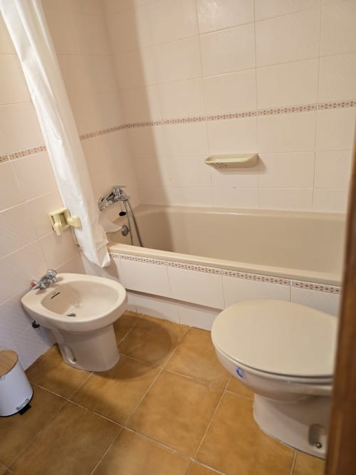 Salle de bain propre avec carrelage blanc, baignoire avec douche, toilettes, bidet et sol en carrelage brun dans un appartement à Roses près de la plage
