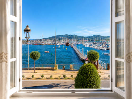 Vista desde ventana abierta de la marina de Baiona con veleros, muelle y montañas bajo cielo azul desde apartamento frente al mar