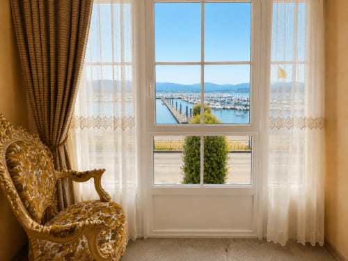 Elegante habitación de hotel en Baiona con vistas al mar de la marina, silla dorada ornamentada y cortinas translúcidas con vistas a la costa.
