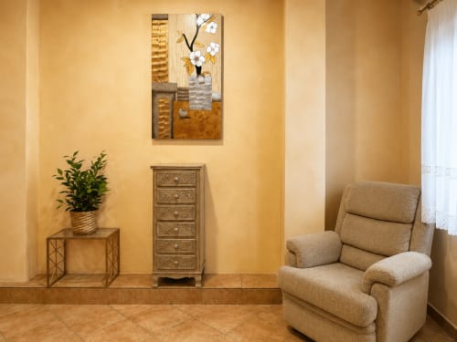 Acogedora sala de estar en Baiona con butaca beige, mesilla de madera con maceta y cuadro floral en paredes amarillas