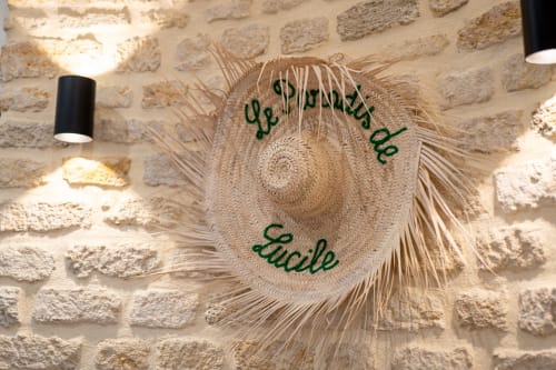 Chapeau de paille avec « Lucile » brodé en vert suspendu au mur de pierre rustique du Paradis de Lucile, La Roche Guyon