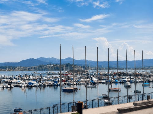 Marina de Baiona con veleros, agua azul, montañas y paseo marítimo con barandilla