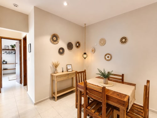 Cálida zona de comedor con mesa de madera, decoración de cestería en la pared y luz natural en la suite del Hotel Isla Mujeres Centro.