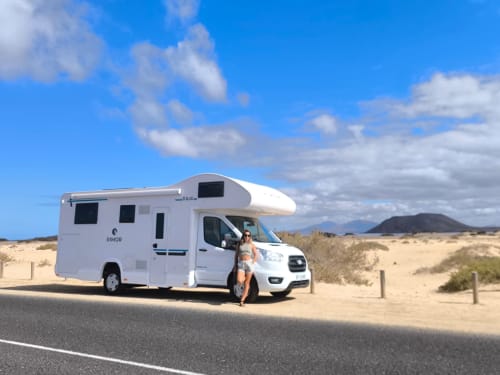 Autocaravana RIMOR blanca estacionada en el paisaje desértico de Fuerteventura con mujer de pie afuera, montañas visibles