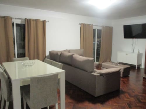 Modern apartment living room at Supranee Apartments with gray sofa, dining table, TV, and beige curtains overlooking Câmara