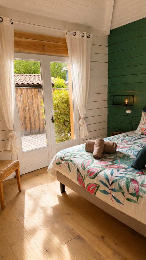 Chambre lumineuse avec mur vert, literie floral et portes vitrées donnant sur terrasse La Loge Du Bassin