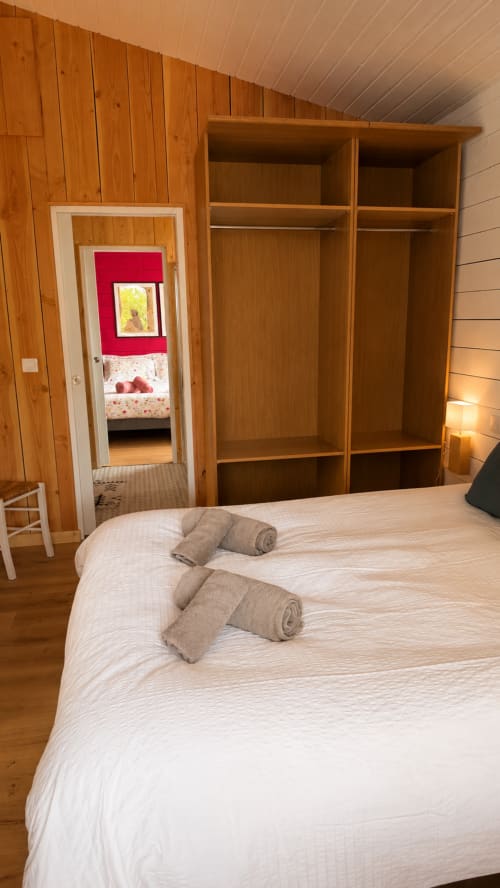 Chambre spacieuse aux poutres bois, grand lit avec serviettes roulées, armoire ouverte, vue sur la chambre d'enfant à Villa La Chunga