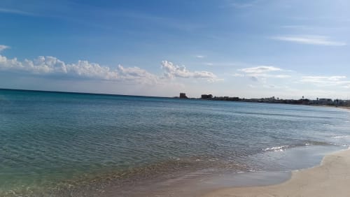 Mare turchese e spiaggia sabbiosa con la cittadina costiera di Veglie visibile sull'orizzonte sotto un cielo azzurro