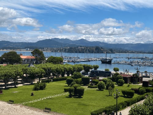 Los jardines del hotel Holibai con vistas a la marina de Baiona, veleros, montañas y paseo marítimo bajo un cielo azul