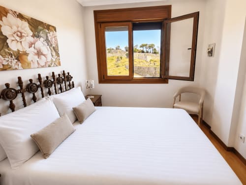 Dormitorio luminoso en el hotel Holibai de Baiona con cama blanca, cabecero de madera y vistas al campo desde grandes ventanales
