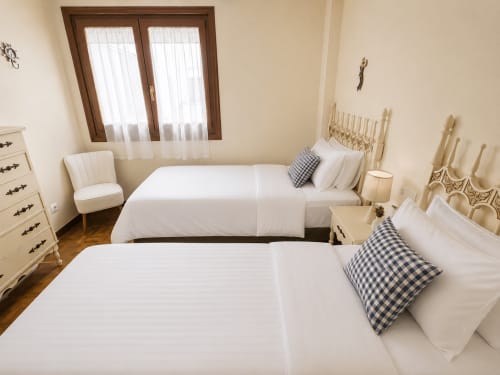 Habitación doble del hotel Holibai en Baiona con ropa de cama blanca, muebles color crema y ventanas de madera