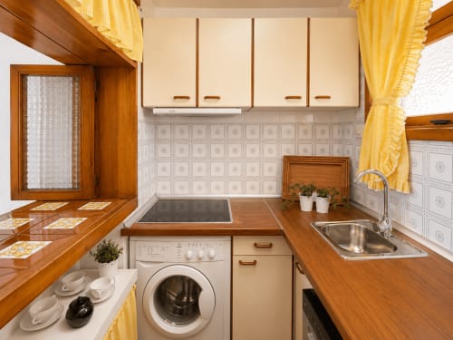 Cocina compacta tipo galley con encimeras de madera, muebles color crema, fregadero de acero inoxidable, lavadora y cortinas amarillas