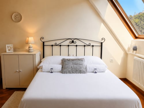 Luminoso dormitorio abuhardillado en Baiona con cama de hierro forjado, ropa blanca, vigas de madera y vistas a la ciudad desde claraboya