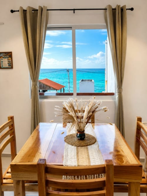 Mesa de comedor de madera con vistas al mar en Hotel Isla Mujeres Centro, cortinas beige enmarcan el océano Caribe turquesa y el cielo