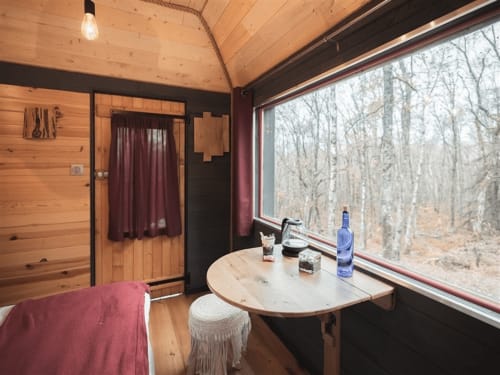 Cozy cabin interior at Les Murmures du Bois with wooden walls, large windows overlooking bare trees, round table with blue