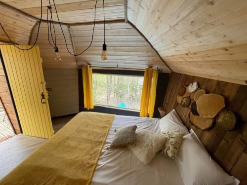 Chambre cosy avec plafond en bois, accents jaunes, lit garni de coussins, grande fenêtre donnant sur le jardin du gîte Les Murmures du Bois