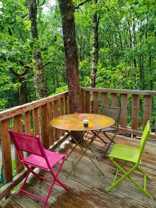 Terrasse en bois avec chaises colorées et table ronde face à la forêt, Gîte Les Murmures du Bois