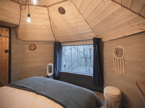 Cozy wooden cabin bedroom with bed, large window overlooking snowy forest, and bohemian decor at Les Murmures du Bois B&B