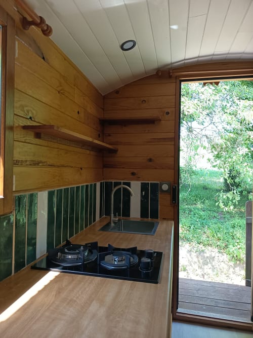 Kitchenette compacte de La Roulotte à Maison Maliga avec murs en bois, cuisinière noire, évier et vue sur le jardin