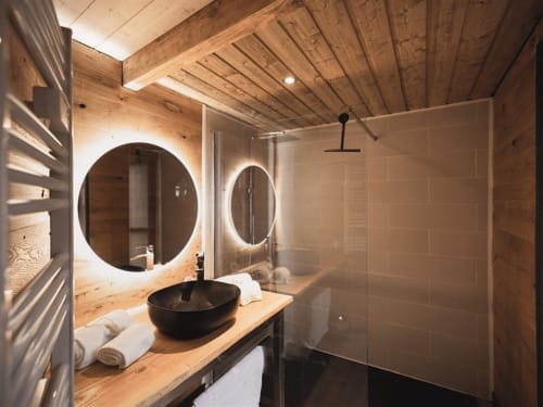 Modern bathroom at Les Murmures du Bois with wooden ceiling, round illuminated mirror, black sink, and walk-in shower