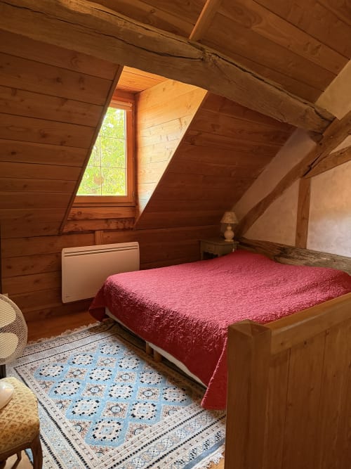 Cozy attic bedroom at Domaine les Bertrands cottage with wooden beams, red bedspread, and garden view window