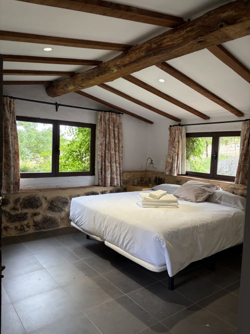 Dormitorio rústico con vigas de madera vista, cama Queen, grandes ventanales y pared de piedra en El Escondite De Pedro Malillo