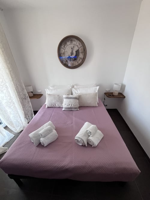 Camera da letto luminosa con biancheria rosa, cuscini bianchi, orologio decorativo e comodini in legno all'hotel Cuore di Rocca