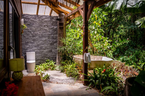 Salle de bains tropicale extérieure du West Indies Cottage en Guadeloupe avec murs en pierre, baignoire, toilettes, végétation luxuriante et boiseries