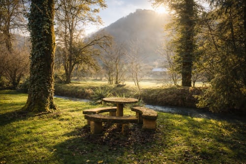 Table de pique-nique en pierre rustique au bord du ruisseau à La Source Bleue, Cusance, entourée d'arbres couverts de lierre et de vues sur la vallée