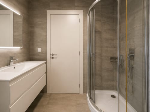 Baño moderno con ducha de cristal curvo, tocador blanco y paredes de azulejos beige en apartamento frente al mar en Nigrán