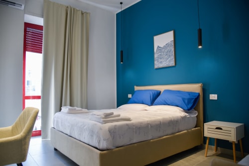 Camera da letto moderna all'Onde Del Mare di Napoli con parete azzurra, letto in legno, cuscini blu e accesso al balcone con vista mare.