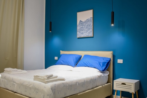 Camera da letto moderna con pareti blu teal intenso, letto bianco, cuscini blu e arredo minimalista all'appartamento Onde Del Mare di Napoli