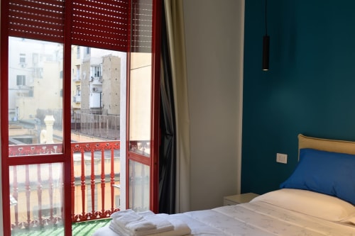 Camera da letto in appartamento napoletano con pareti turchesi, letto in legno, cuscino blu e porta-balcone con cornice rossa che affaccia su edifici cittadini